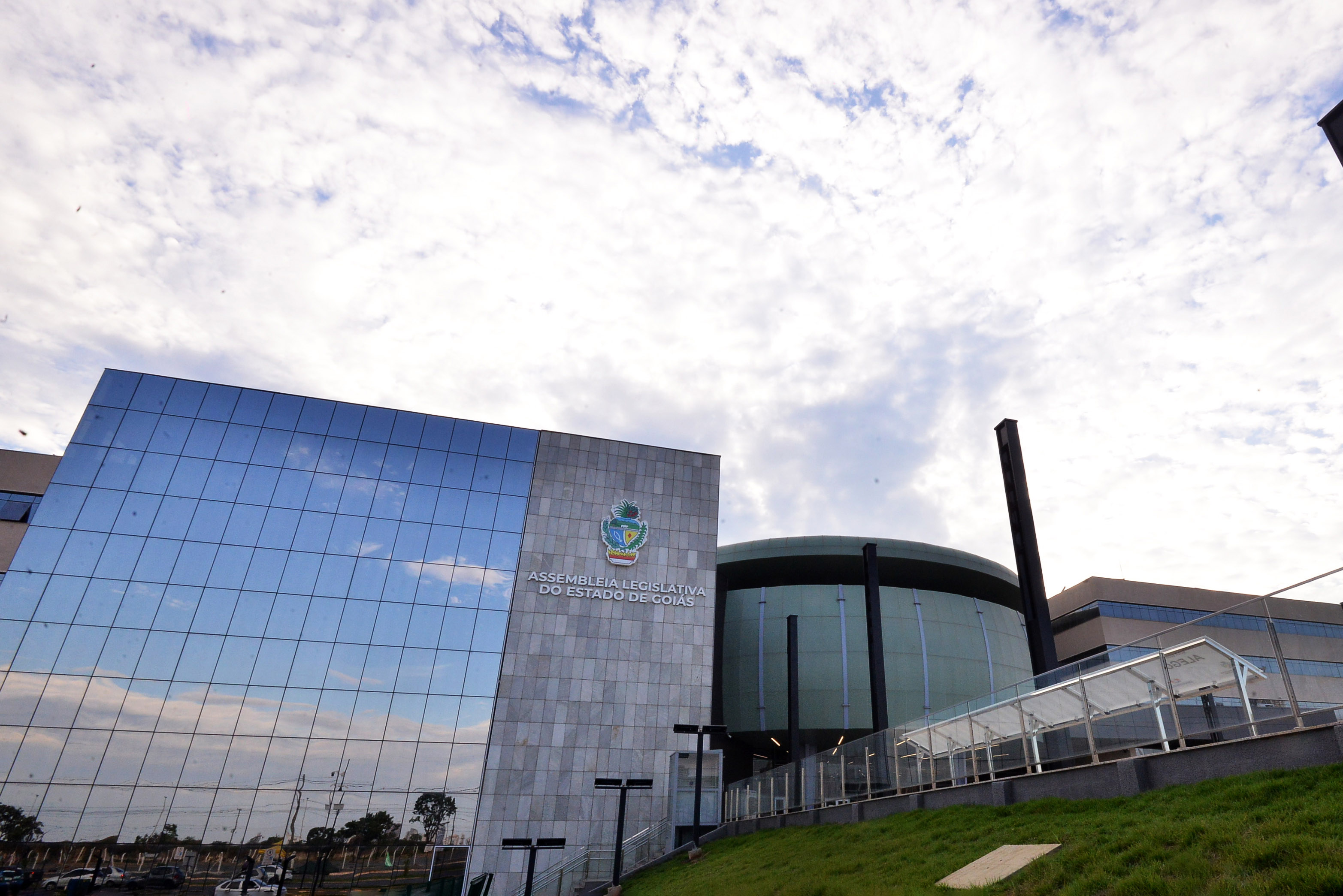 Palácio Maguito Vilela, sede da Assembleia Legislativa de Goiás, visto em perspectiva: fachada de vidro, bloco de pedra com o brasão e o volume arredondado da entrada sob céu com nuvens