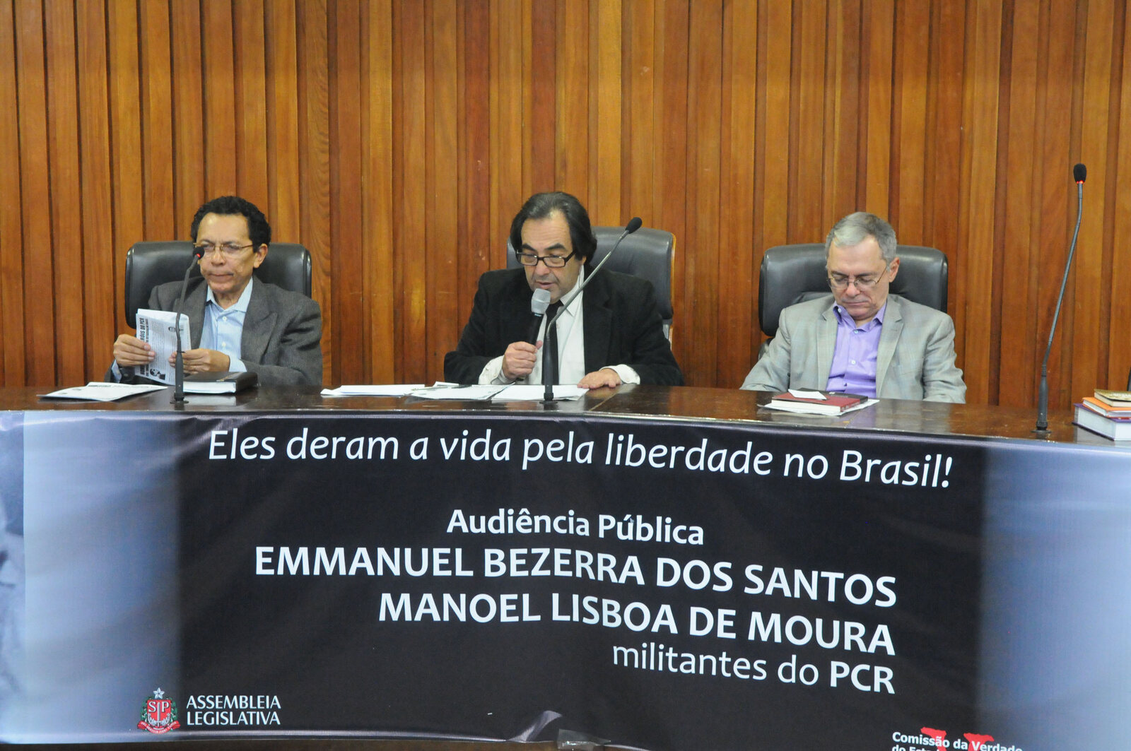 Três homens sentados à mesa de uma audiência pública na Alesp, diante de painel de madeira, um deles falando ao microfone