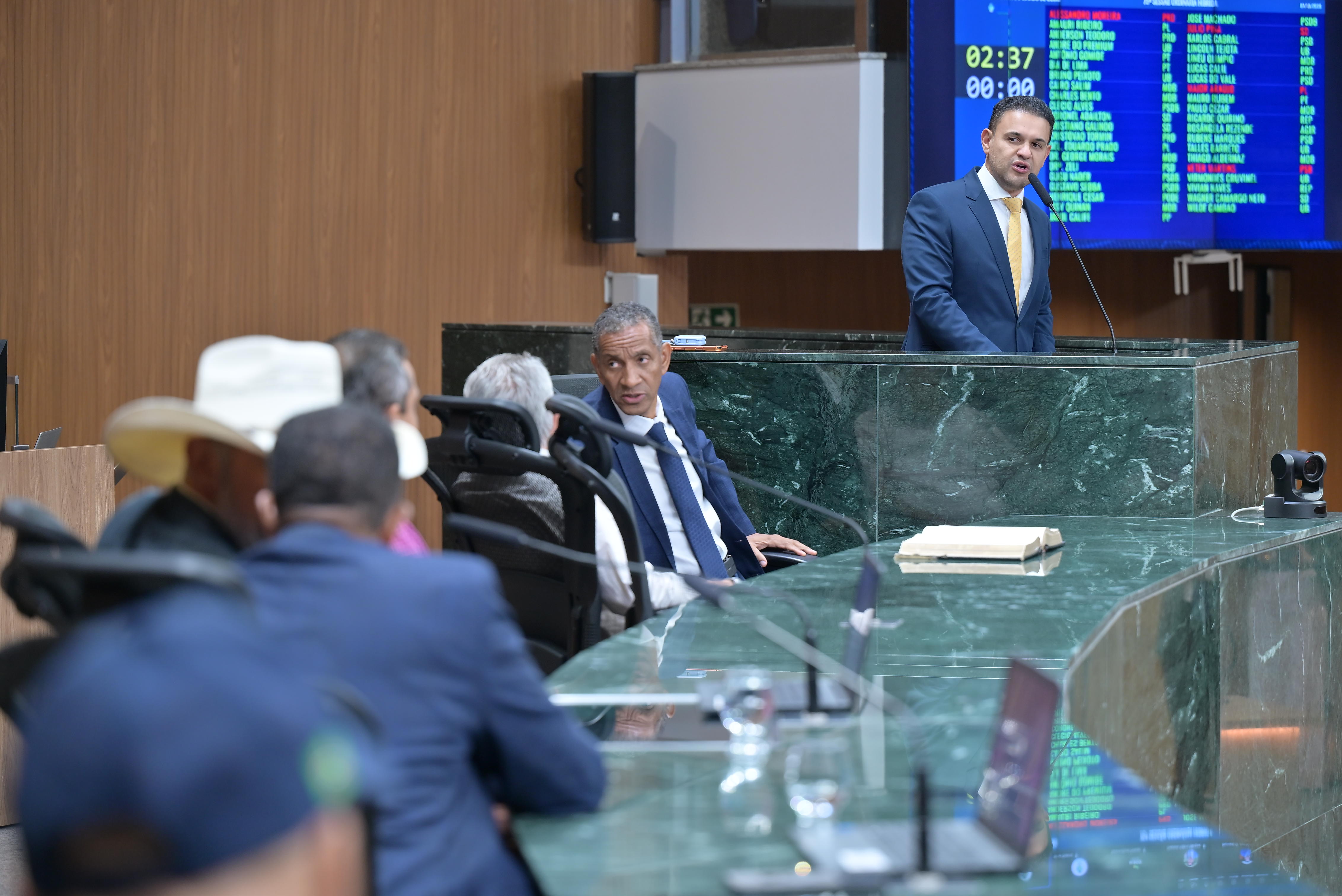 Vista lateral do plenário da Assembleia Legislativa de Goiás durante sessão, com deputados nas bancadas vistos de costas em primeiro plano e um deputado discursando na tribuna ao fundo