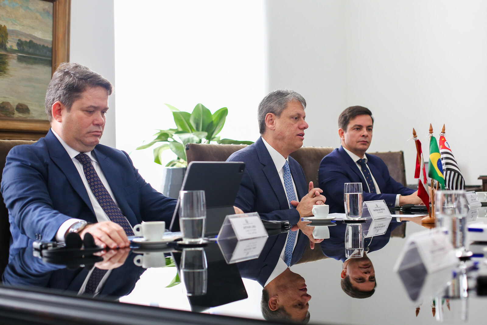 Tarcísio de Freitas fala sentado a uma mesa de reunião, entre dois homens de terno, no Palácio dos Bandeirantes