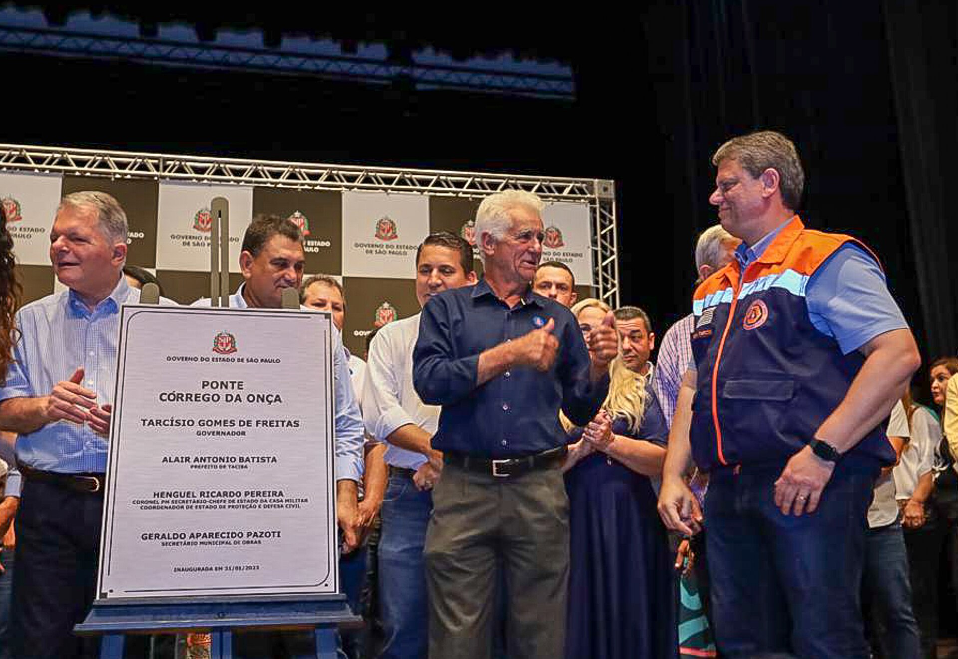 Tarcísio de Freitas, de colete da Defesa Civil, aplaude ao lado de prefeitos e moradores diante de placa de inauguração
