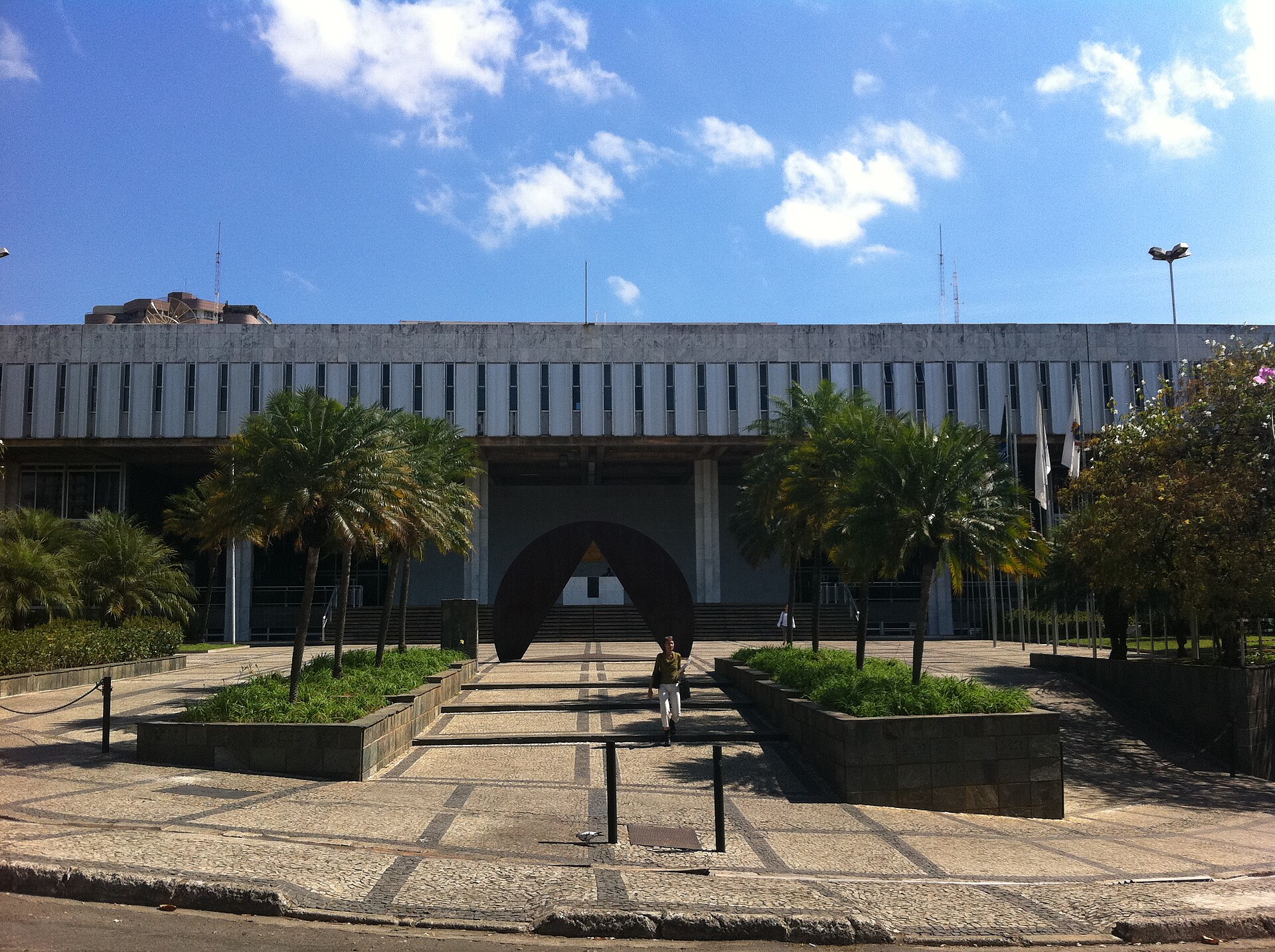 Fachada da Assembleia Legislativa de Minas Gerais, com palmeiras e jardins diante do prédio, sob céu azul