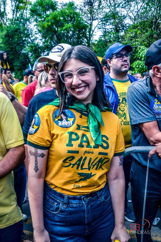Eduarda Campopiano sorri de óculos, camiseta amarela e lenço verde no pescoço, entre apoiadores em carreata ao ar livre