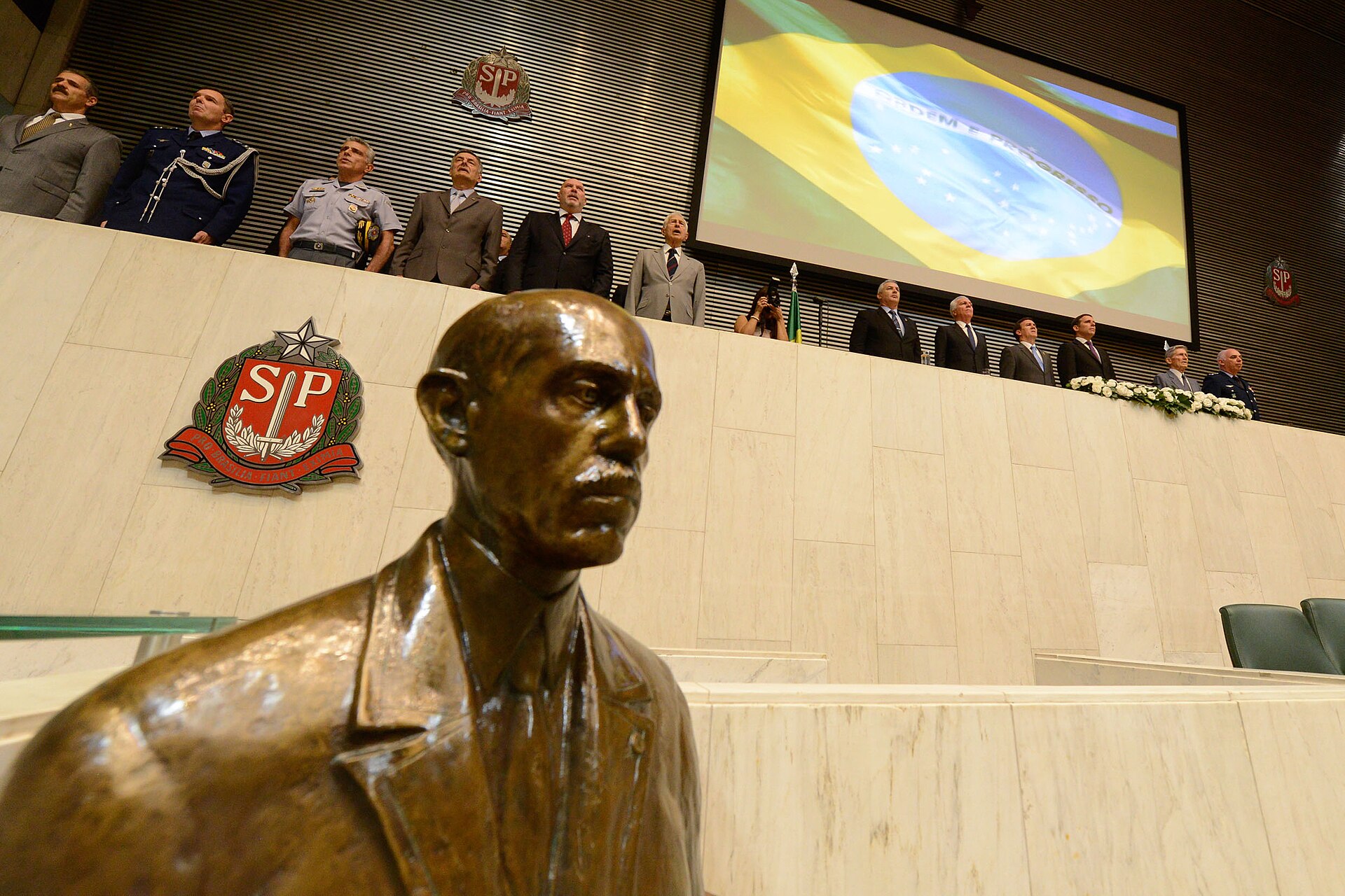 Plenário da Alesp visto de ângulo lateral, com Mesa ao fundo e busto de bronze em primeiro plano