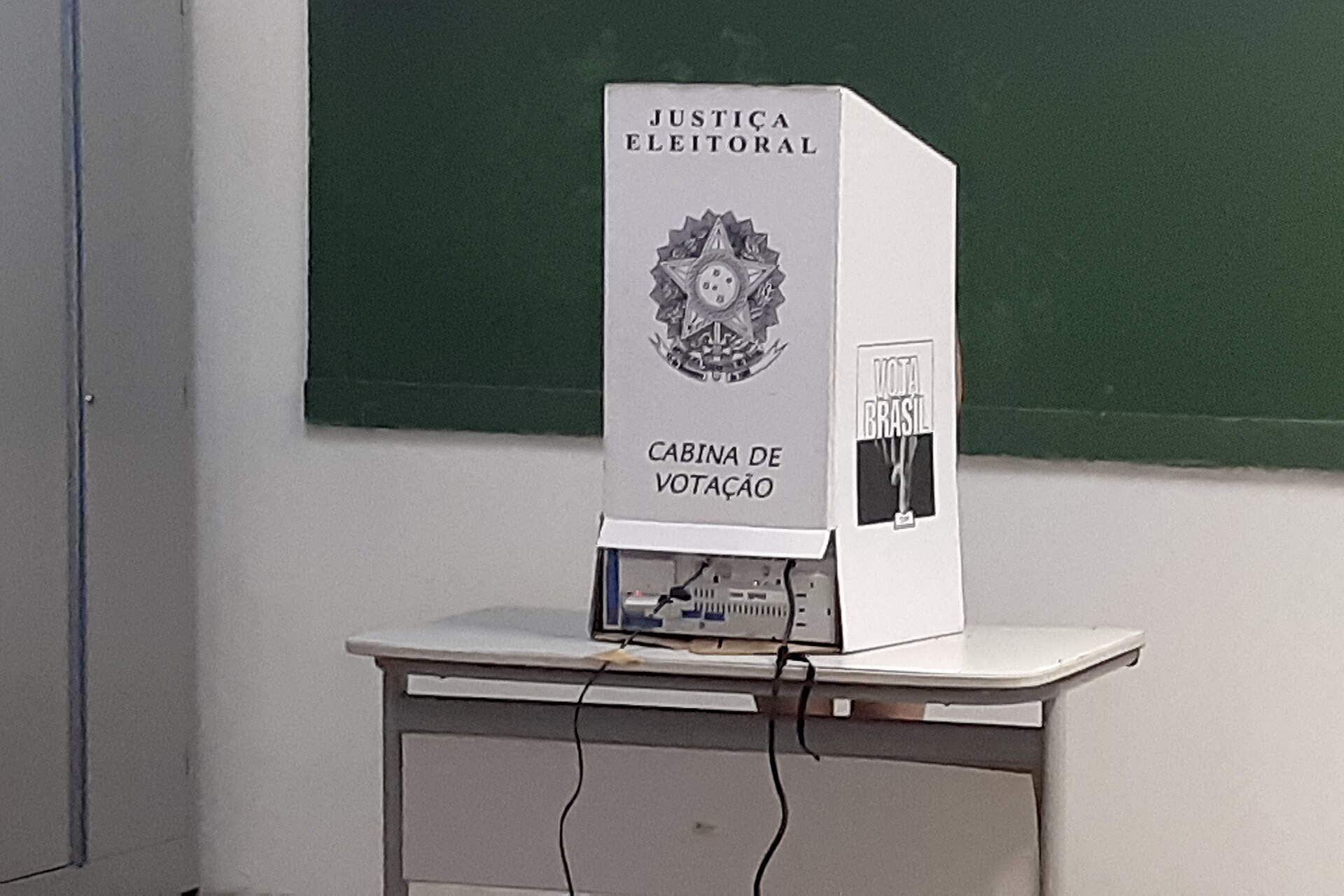 Cabine de votação da Justiça Eleitoral, com a urna eletrônica sobre uma carteira escolar, sem nenhum eleitor diante dela