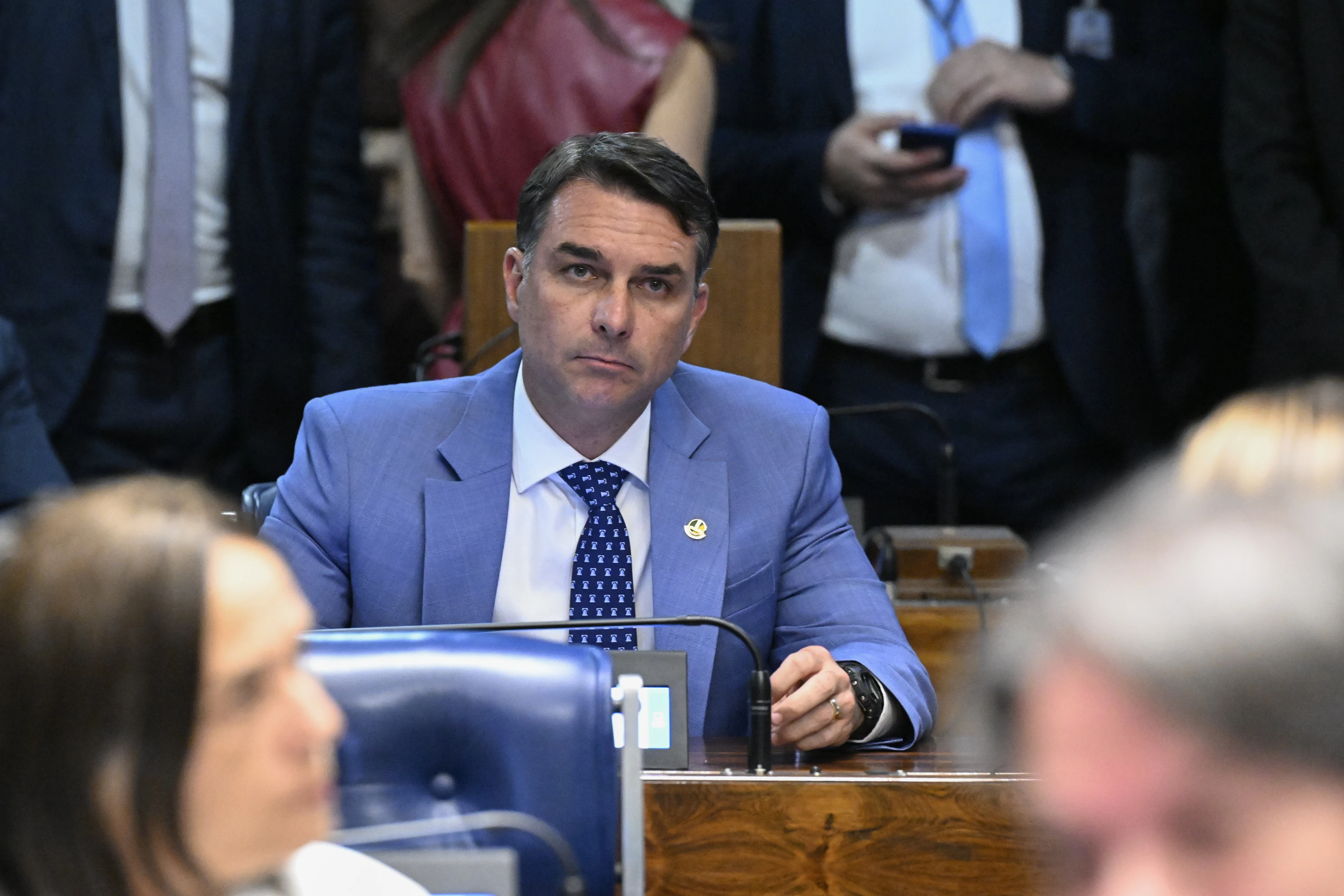 Flávio Bolsonaro de terno azul à bancada do plenário do Senado, com o rosto em destaque e um microfone à frente