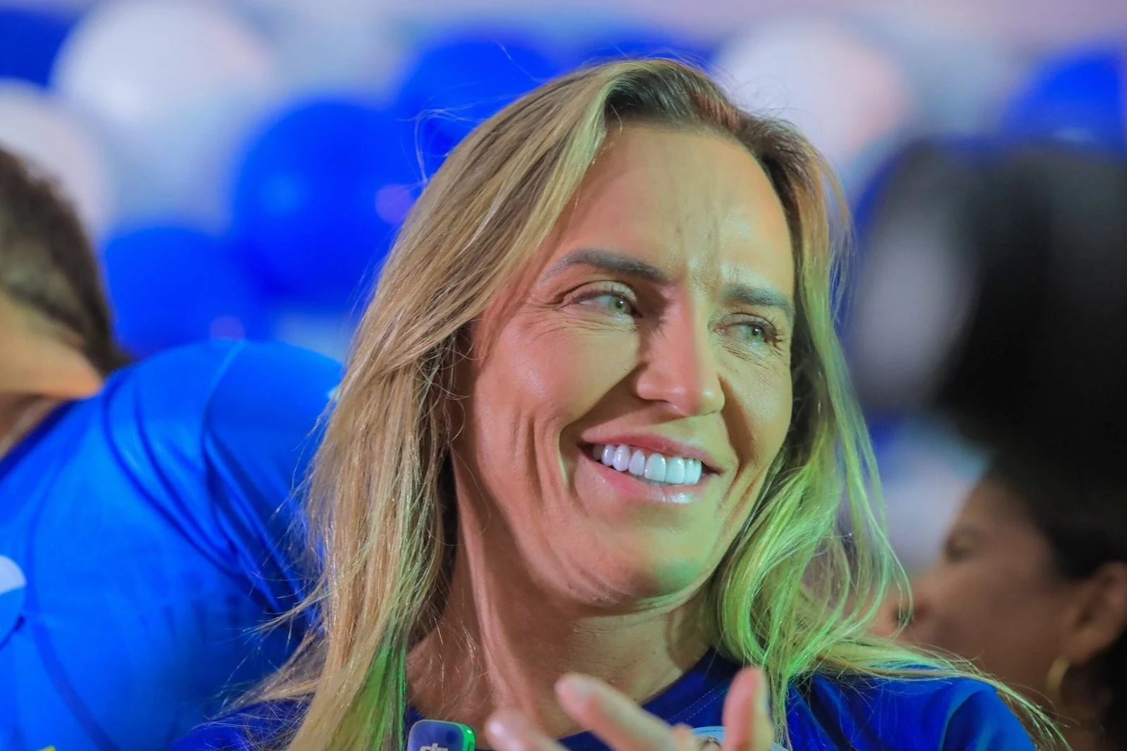 Celina Leão em close, sorrindo com camisa azul entre apoiadores, com o rosto em destaque