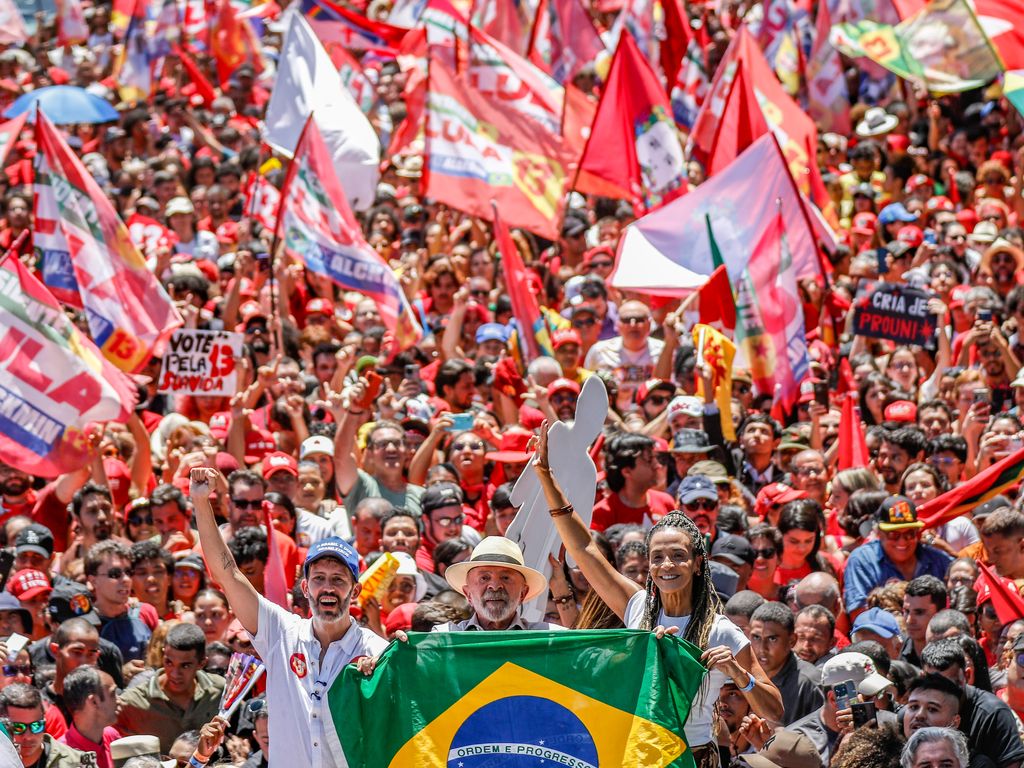 Lula, de chapéu, sorri entre apoiadores que erguem uma bandeira do Brasil diante de uma multidão com bandeiras vermelhas