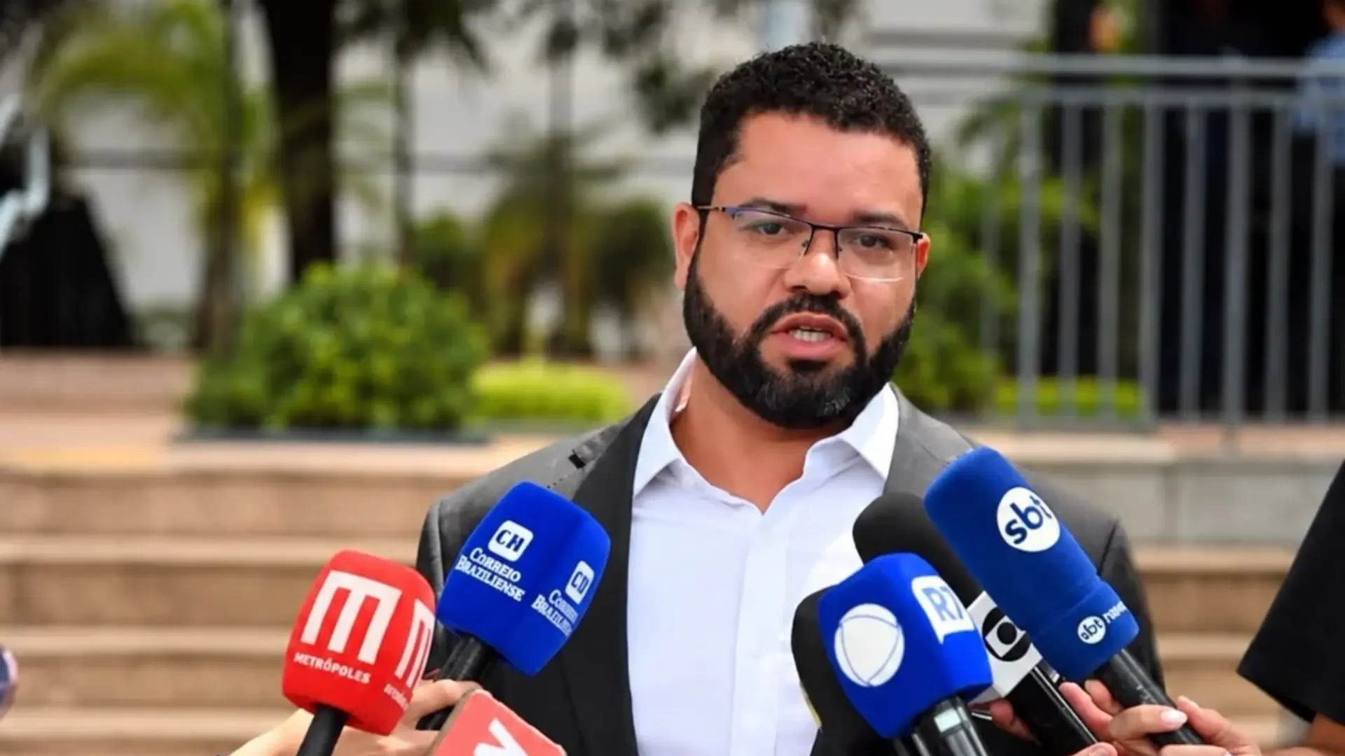 Elisson Ferreira, de óculos e paletó cinza, fala diante de microfones de emissoras ao ar livre.