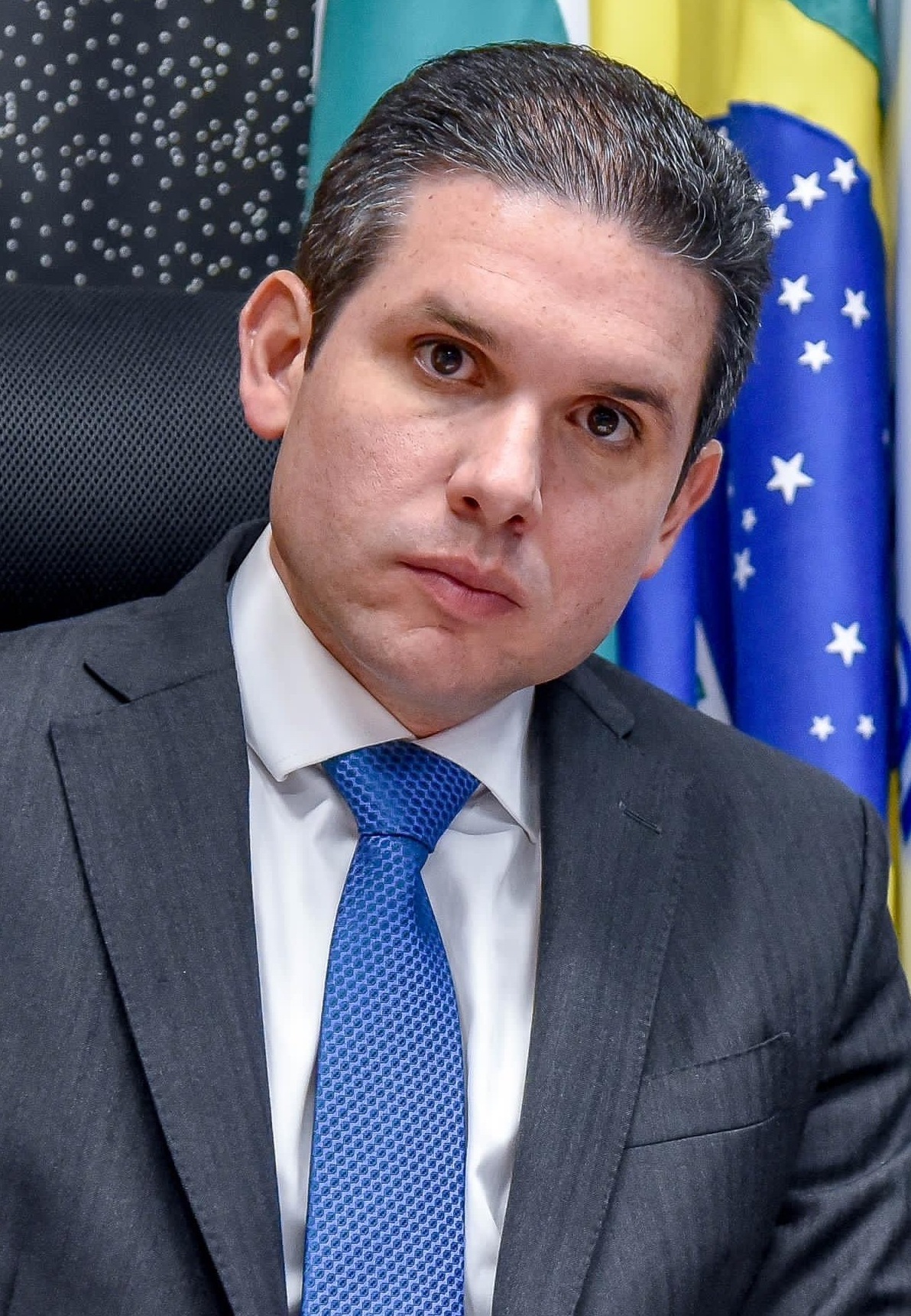 Hugo Motta em close, de terno cinza-escuro e gravata azul, com bandeira do Brasil ao fundo